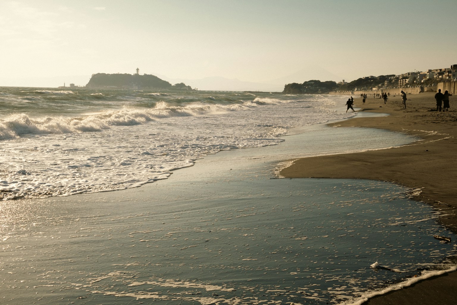 Waves gently lap a sandy beach with distant people. (Photo by Kate Kasiutich on Unsplash)