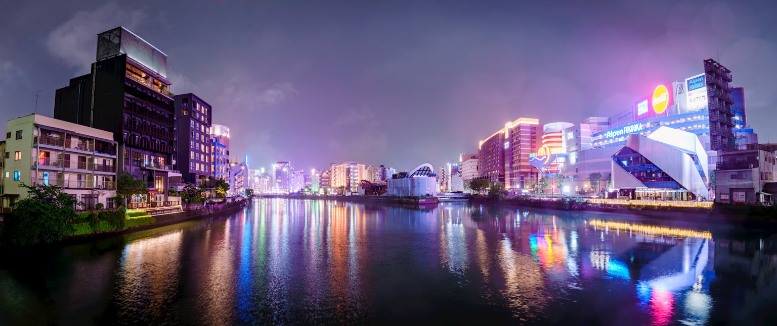 Cityscape with illuminated buildings reflecting in water at night (Photo by Jayjayli on Unsplash)